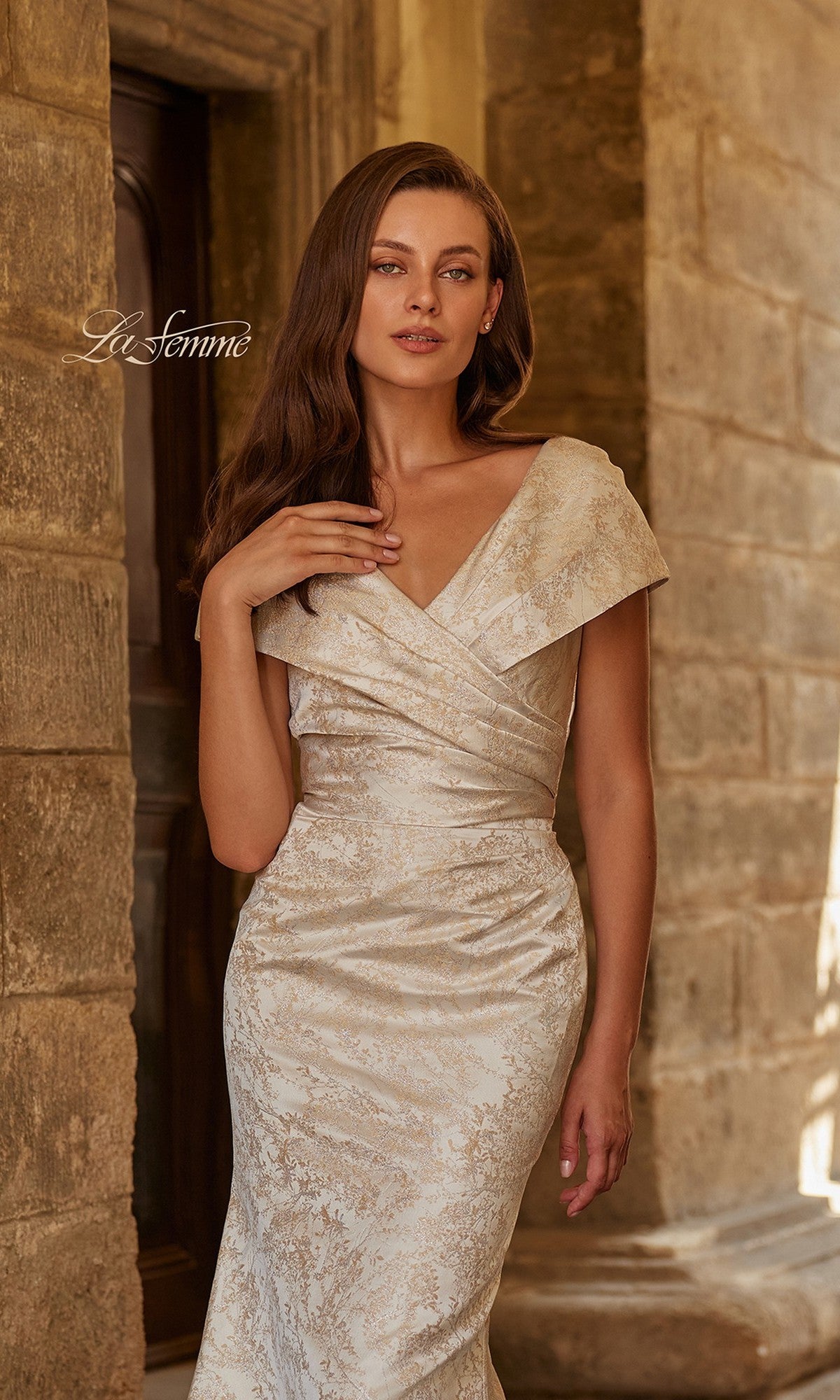 A woman wearing the Long Formal Dress: La Femme 33428, an off-shoulder ivory jacquard evening gown, stands against a stone wall and gazes at the camera with one hand gently touching her chest.